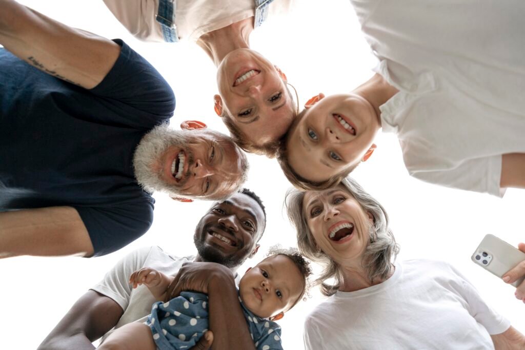Grupo multigeracional e diverso formado por crianças, adultos e idosos sorrindo enquanto olham para a câmera posicionada abaixo deles. A imagem representa diferentes gerações reunidas em um momento de convivência, simbolizando mudanças comportamentais, conexões familiares, diversidade etária e transformações sociais após o período de quarentena.