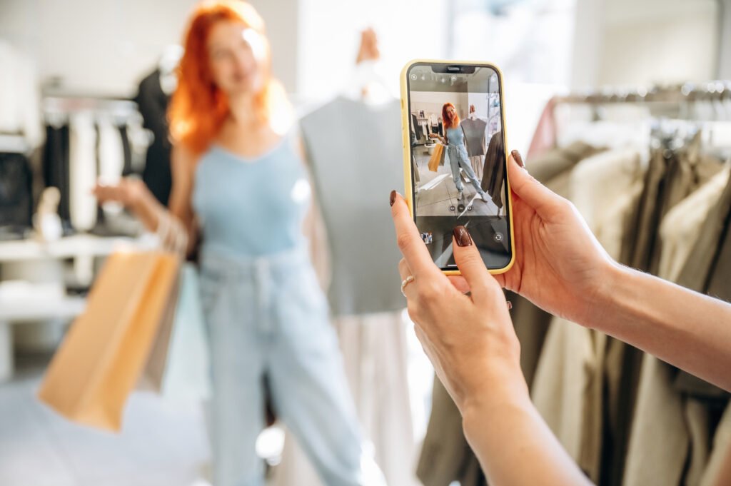 Pessoa fotografando uma mulher em uma loja de roupas usando um smartphone. A mulher posa segurando uma peça de roupa e sacolas de compras, enquanto sua imagem aparece na tela do celular. A cena representa criação de conteúdo para redes sociais, marketing digital, influência digital e o uso do Instagram Shopping para divulgar e vender produtos de moda online.