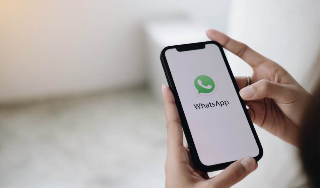 Pessoa segurando um smartphone com a tela exibindo o logotipo do WhatsApp em destaque. A imagem representa pagamentos e transações comerciais realizadas pelo aplicativo de mensagens, associando praticidade, comunicação instantânea e facilidades para empresas no atendimento e recebimento de clientes.