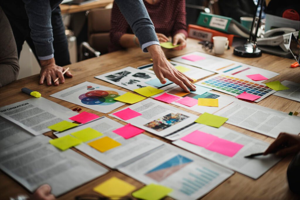 Equipe reunida em torno de uma mesa analisando documentos, gráficos, amostras de cores e post-its coloridos durante uma sessão de planejamento e mapeamento estratégico.