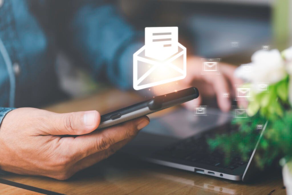 Pessoa segurando um smartphone enquanto utiliza um notebook sobre uma mesa. Acima do celular, ícones de envelopes representam o envio e recebimento de e-mails, simbolizando newsletters, comunicação digital e estratégias de e-mail marketing.