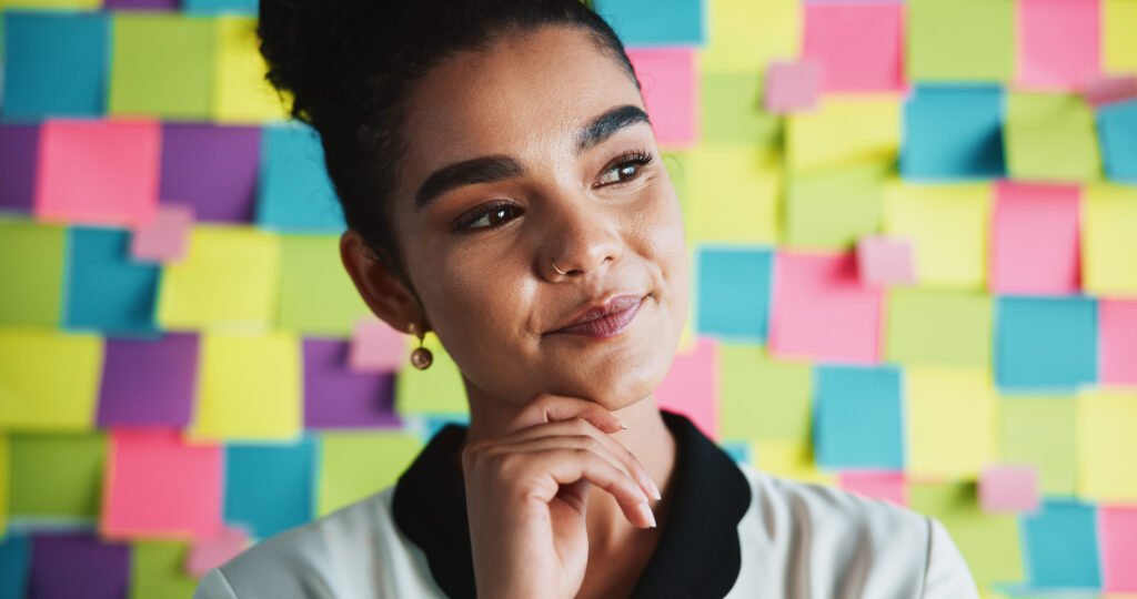 Mulher em primeiro plano, com expressão pensativa e mão apoiada no queixo, diante de uma parede coberta por post-its coloridos, representando planejamento estratégico, criatividade e construção de identidade de marca.