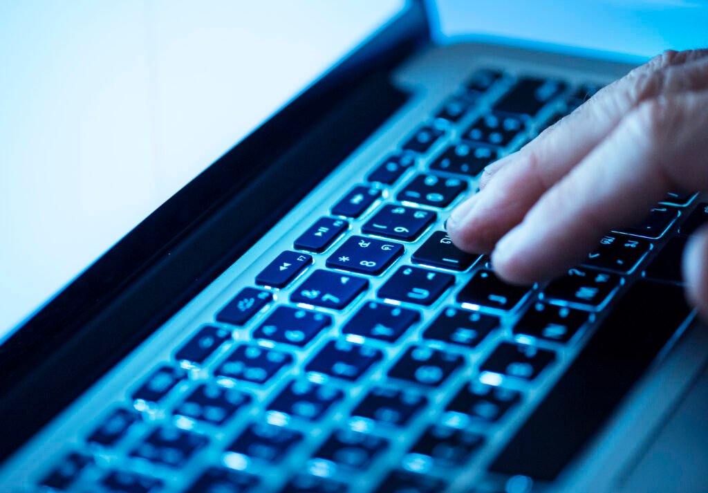 Close-up de uma mão digitando em um teclado de notebook iluminado por luz azul. Os dedos pressionam as teclas enquanto o restante do teclado aparece desfocado, criando uma sensação de tecnologia, produtividade e trabalho digital.