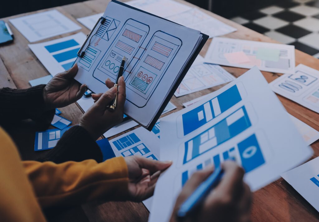 Profissionais analisando wireframes e protótipos de interfaces mobile impressos sobre uma mesa, discutindo a estrutura e a organização de elementos da experiência do usuário para aplicativos e produtos digitais.