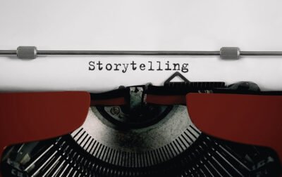 Máquina de escrever antiga com a palavra “Storytelling” digitada em uma folha de papel branca. A imagem destaca a fita vermelha e os mecanismos metálicos da máquina, simbolizando a criação de narrativas para engajar e conectar marcas ao público.