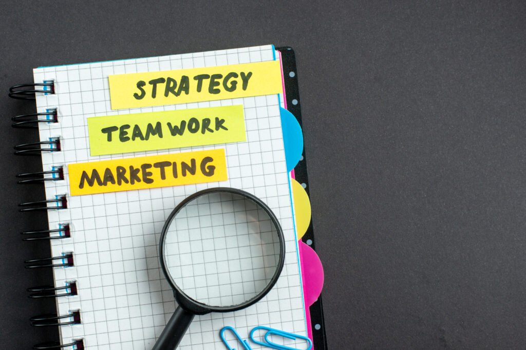 Caderno aberto com anotações relacionadas a marketing, estratégia e trabalho em equipe, acompanhado por uma lupa sobre a página. Etiquetas coloridas destacam os termos “Strategy”, “Team Work” e “Marketing”, representando planejamento, análise e ações voltadas ao engajamento e ao fortalecimento da presença digital nas redes sociais.