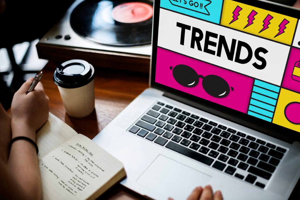 Pessoa analisando tendências digitais em um notebook com a palavra “Trends” destacada na tela, enquanto faz anotações em um caderno. Ao lado, há um copo de café e um toca-discos, simbolizando pesquisa de mercado, monitoramento de tendências e planejamento estratégico.