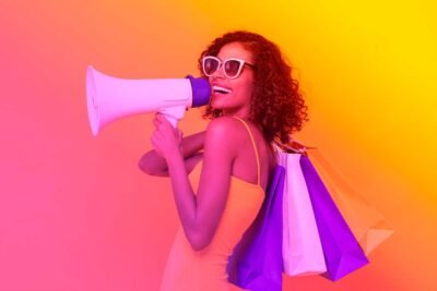 Mulher segurando sacolas coloridas e megafone representa ação de marketing com impacto visual e entusiasmo.