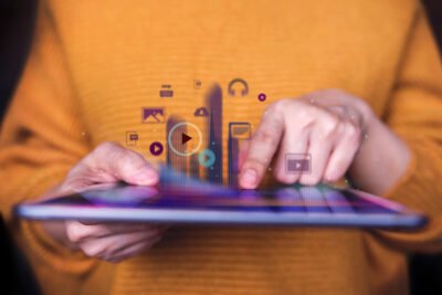 Pessoa utilizando um tablet enquanto ícones holográficos de vídeos, imagens, áudio e mídias digitais aparecem sobre a tela, simbolizando gestão de conteúdo e tecnologia digital.